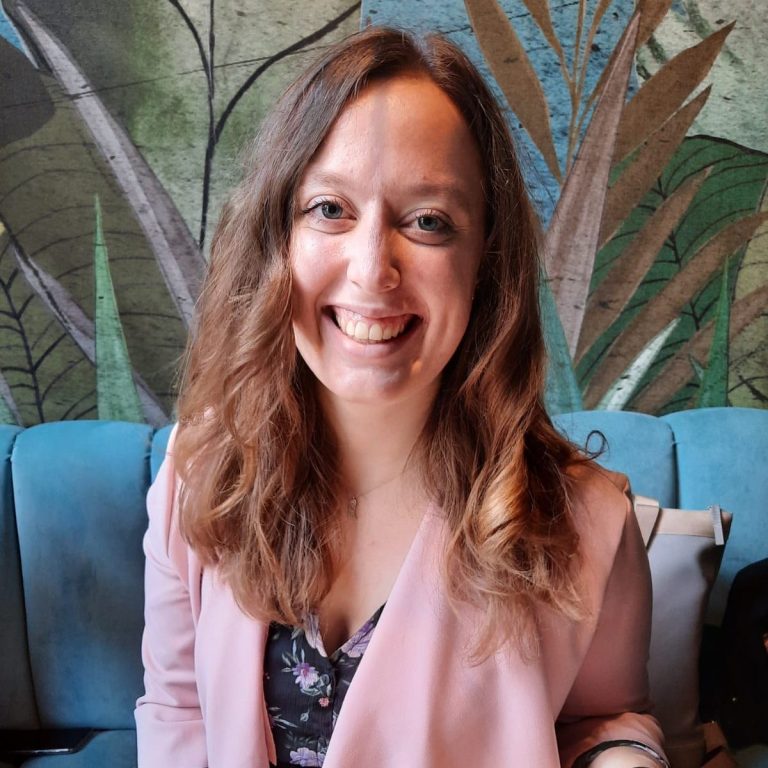 Sofia Lächelnde Frau mit langen, lockigen Haaren, in einer hellen Umgebung mit Pflanzenmotiven.